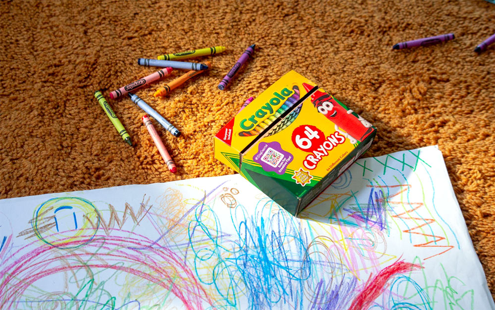 Crayola 64 count box of crayons atop a child's crayon drawing sprawled on a carpeted floor